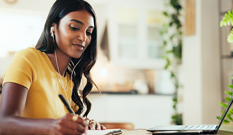 Professional black woman wearing gold shirt, sitting in front of laptop and taking notes with pen and paper